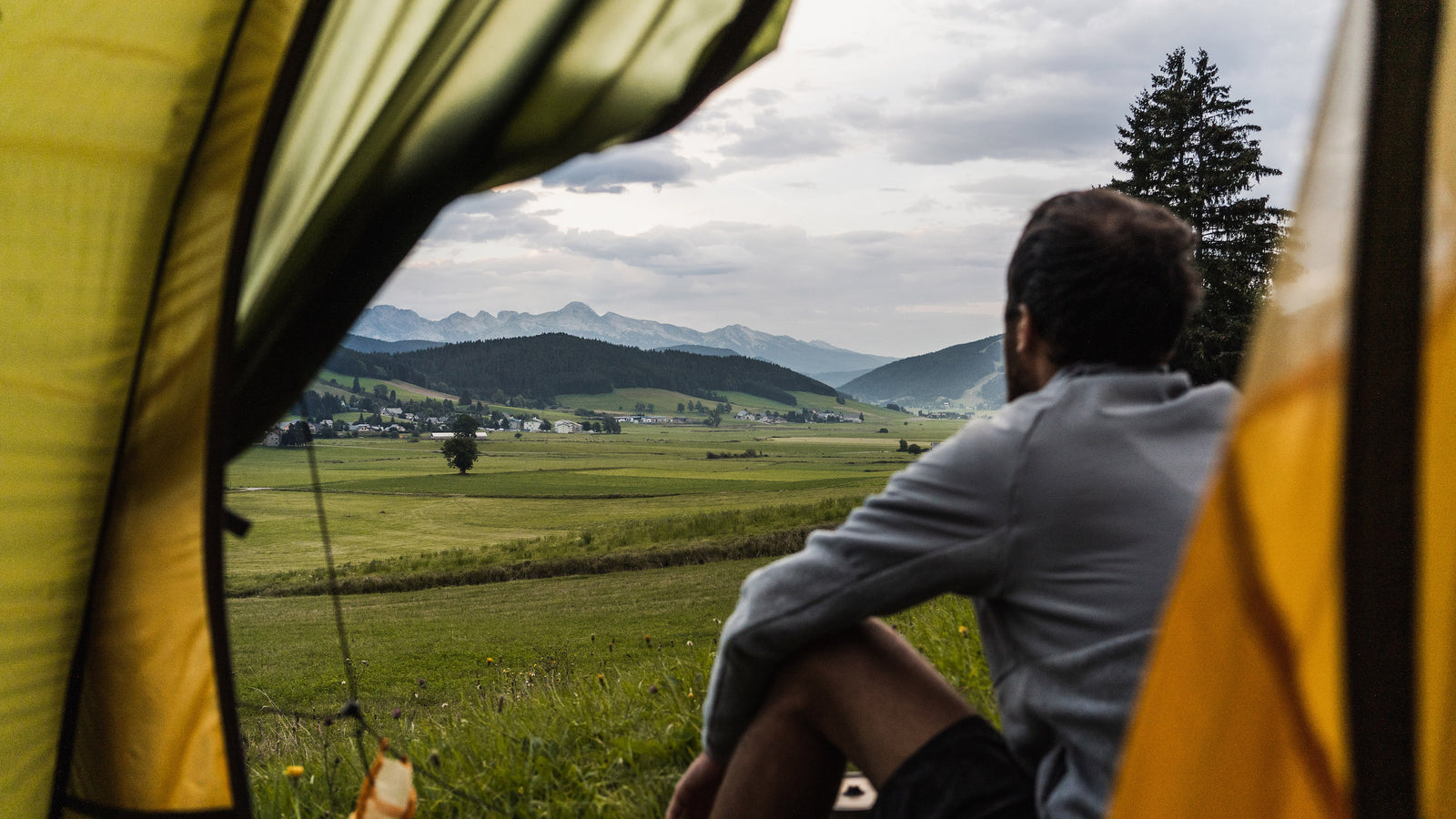 Eine Person die vor einem geöffnetem Zelt sitz und auf die Grüne Landschaft schaut.